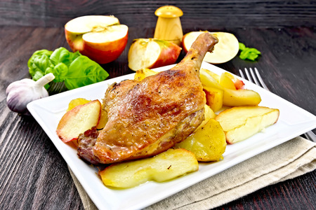 Roasted duck leg with apple, potatoes in a plate on a napkin, basil, garlic and fork on a dark wooden boardの写真素材