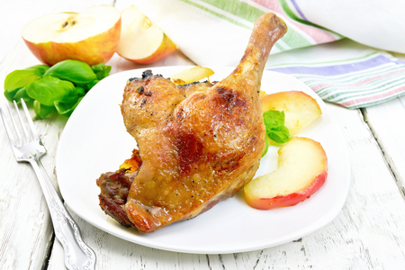 Roasted duck leg with apple, potatoes in a white plate, basil, fork and a kitchen towel on the background light wooden boardsの写真素材