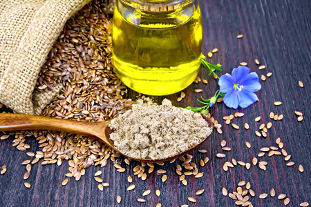 Flaxseed flour in a spoon, oil in a glass jar, blue linen flower and brown seeds in a bag on a wooden board backgroundの写真素材
