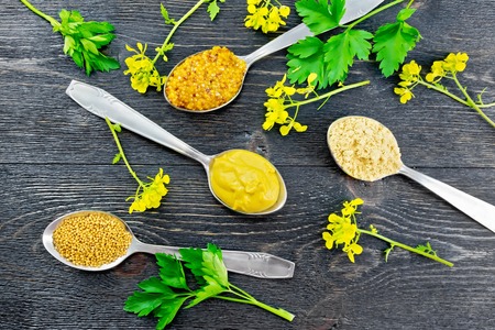 Mustard grainy, powder, seeds and sauce in spoons, yellow mustard flowers and parsley on the background of a wooden board from aboveの写真素材