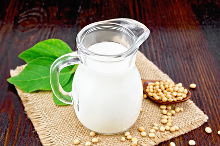 Soy milk in a jug, green leaf, soybeans in a spoon and on sackcloth against a dark wooden boardの写真素材