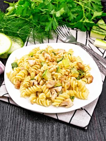 Fusilli pasta with chicken breast, zucchini, cream and pine nuts in a white plate on towel, garlic, fork and parsley on a dark wooden board backgroundの写真素材