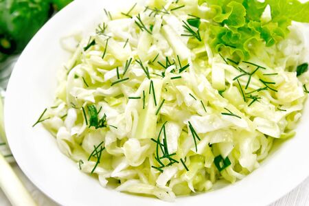 Salad of fresh cabbage, green onions and cucumber with vinegar and vegetable oil dressing in a plate, towel, dill, lettuce and fork on wooden board backgroundの写真素材