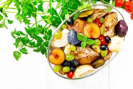 Baked chicken with tomatoes, apples, plums and grapes in a glass roaster on a kitchen towel, garlic, parsley and basil on background of light wooden board from aboveの写真素材