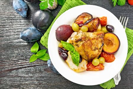 Baked chicken with tomatoes, apples, plums and grapes in a plate on a napkin, garlic, parsley and basil on wooden board background from aboveの写真素材
