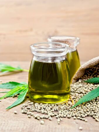 Hemp oil in two glass jars with grain in the bag and on the table, cannabis leaves on the background of wooden boardsの写真素材