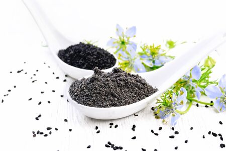 Flour and seeds of Nigella sativa in two spoons, sprigs of kalingini with blue flowers and green leaves on a wooden board backgroundの写真素材