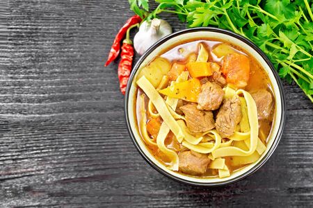 Central Asian national dish Lagman of meat, noodles and vegetables in bowl on a napkin, garlic, parsley on black wooden board from aboveの写真素材