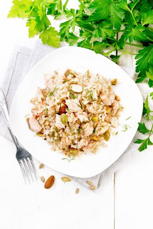 Salad of smoked salmon, rice, sunflower and pumpkin seeds, almonds, seasoned with honey and olive oil in a plate on a towel, parsley and fork on background of light wooden board on topの写真素材