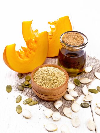 Pumpkin flour in a bowl and oil in a jar on burlap, seeds on table and slices of vegetable on  light wooden boardの写真素材