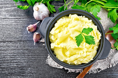 Mashed potatoes in a black saucepan and a spoon on burlap, garlic, parsley on wooden board...