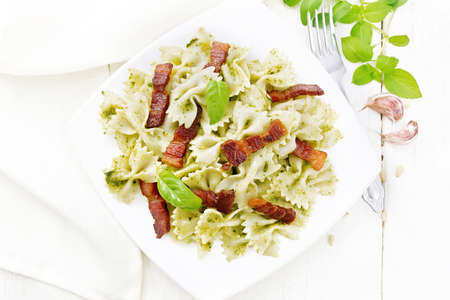 Farfalle pasta with pesto sauce, fried bacon and basil in a plate, garlic, fork and a napkin on light wooden board background from aboveの写真素材