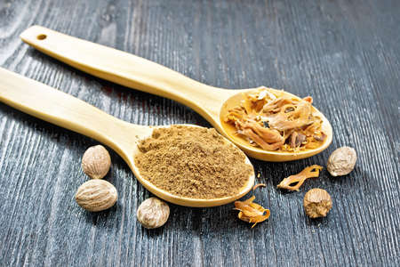 Ground nutmeg and dried nutmeg arillus in two spoons, whole nuts on a wooden board background