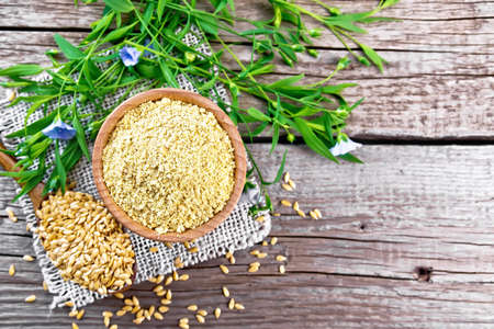 Linen bran in a bowl, seeds in a spoon on burlap napkin, flax leaves and flowers on the background of an old wooden board from aboveの写真素材