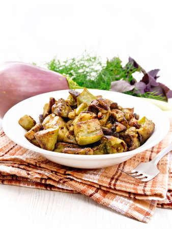 Eggplant fried with onion, egg and spices in a plate on a napkin, basil, leek and dill on light wooden board backgroundの写真素材