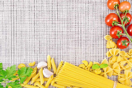 Frame made of different pasta, tomatoes, garlic and parsley on a background of wicker fabricの写真素材