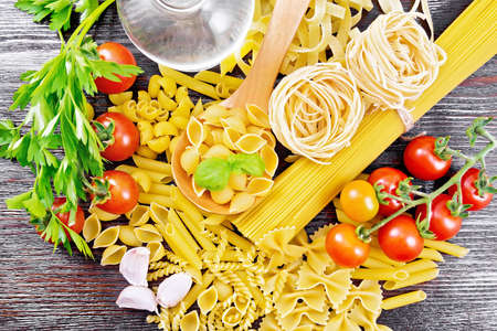 Pasta conchiglie in spoon, pipe rigate, spaghetti, tagliatelle, fusilli, penne, farfalle, conquilla, trenette, rotini, tomatoes, oil in decanter and parsley on wooden board background from aboveの写真素材