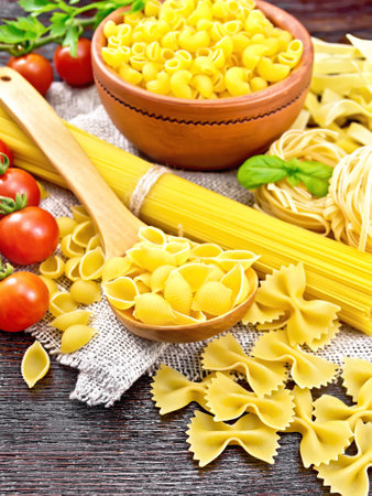Pasta seashells in spoon, pipe rigate in a bowl, spaghetti, tagliatelle, farfalle, trenette, tomatoes and parsley on burlap on wooden board backgroundの写真素材