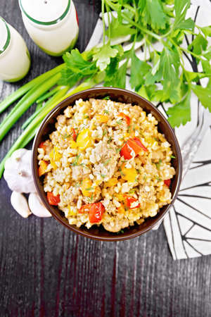 Pearl barley porridge with minced meat, tomato, yellow bell pepper, garlic and onions in a clay bowl, a towel and parsley on dark wooden board background from aboveの写真素材
