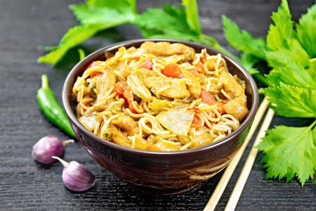 Noodles with cabbage, chicken breast, sweet red peppers, onions and peas, seasoned with soy sauce in a bowl, garlic and green celery leaves on wooden board backgroundの写真素材