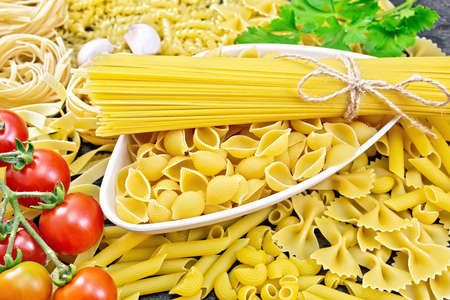 Pasta seashells and spaghetti in bowl, pipe rigate, tagliatelle, fusilli, penne, farfalle, conquilla, trenette, rotini, pappardelle, tomatoes, garlic and parsley on wooden board backgroundの写真素材