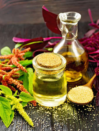 Amaranth oil in a glass jar and decanter, amaranth seeds, brown, green and purple flowers and plant leaves on dark wooden board backgroundの写真素材