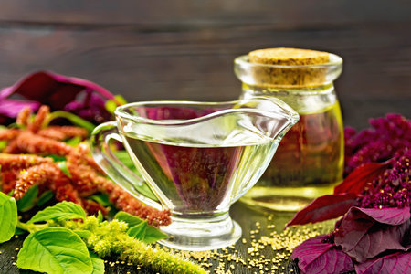 Amaranth oil in a glass gravy boat and jar, brown, green and purple flowers and leaves of the plant on a dark wooden board backgroundの写真素材