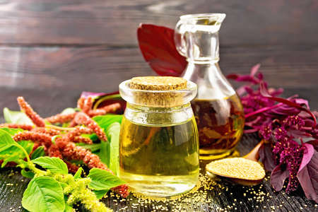 Amaranth oil in a glass jar and decanter, amaranth seeds in a spoon, brown, green and purple flowers and plant leaves on a wooden board backgroundの写真素材