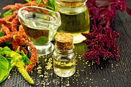 Amaranth oil in a glass bottle, gravy boat and jar, brown, green and purple flowers and leaves of the plant on the background of wooden boardの写真素材