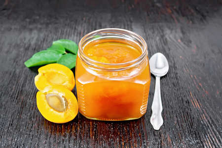 Jam apricot in glass jar, fruits, a spoon and a branch with green leaves on the background of dark wooden boardの写真素材