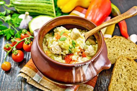 Rice with chicken, bell pepper, zucchini and carrots in a clay pot with a spoon on towel, bread and vegetables on wooden board backgroundの写真素材