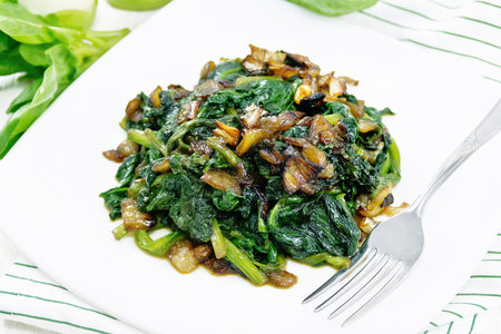 Spinach fried with onions in a plate on a napkin on background of light wooden boardの写真素材