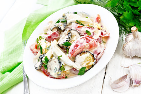 Salad with fried zucchini, boiled egg, fresh tomato and garlic, dressed with mayonnaise or sour cream in a plate, a towel and parsley on the background of light wooden boardの写真素材
