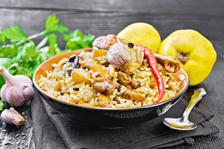 Pilaf from rice, duck, quince, carrots with garlic, onion, prunes and spices in a plate on a towel against the background of dark wooden boardの写真素材
