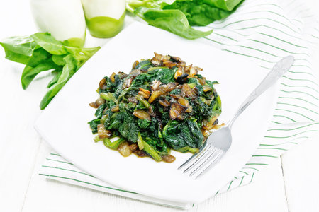 Spinach fried with onions in a plate on a towel against the background of white wooden boardの写真素材