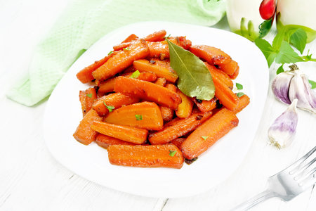 Carrots fried with garlic, bay leaf, spices and green onions in a plate, basil and a napkin on wooden board backgroundの写真素材