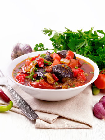 Vegetable ragout with eggplant, tomatoes, bell peppers, onions and spices in a plate on napkin, garlic, parsley, hot peppers and a fork on wooden board backgroundの写真素材