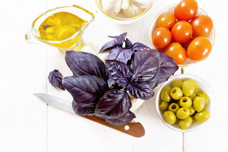 Fresh purple basil in a mortar, olives, tomatoes and champignons in bowls, vegetable oil in gravy boat and a knife on light wooden board background from aboveの写真素材