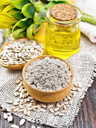 Sunflower flour in bowl, oil in glass jar, seeds in a spoon on a burlap napkin, sunflower flower bud on dark wooden board backgroundの写真素材