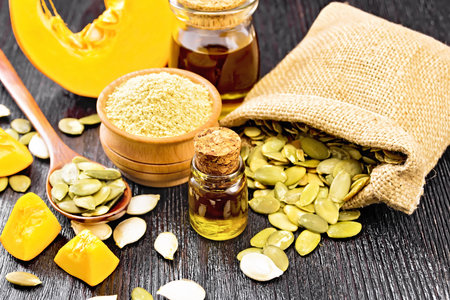 Pumpkin oil in vial and a jar, flour in a bowl, seeds in a spoon and bag, slices of vegetable on dark wooden board backgroundの写真素材
