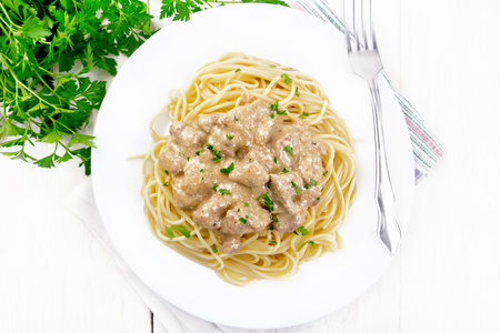 Pieces of meat in creamy sauce with spaghetti in a plate on a napkin on background of a wooden board from aboveの写真素材
