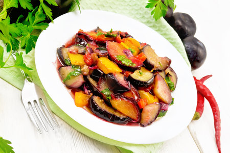 Eggplants fried with plums, onions and sweet peppers in a plate on a napkin against the background of wooden boardの写真素材