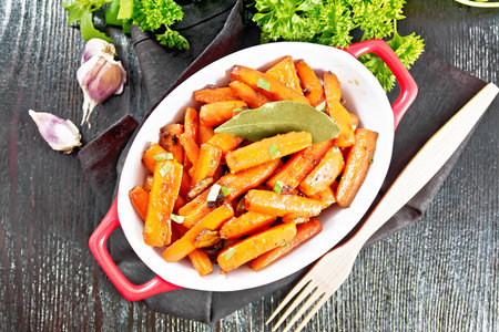 Carrots fried with garlic, bay leaf, spices and green onions in a pan on a napkin, basil and parsley on wooden board background from aboveの写真素材