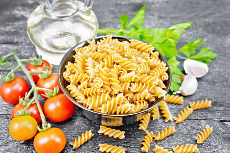 Whole grain wheat flour fusilli pasta in a bowl, tomatoes, garlic, vegetable oil in a decanter and parsley on dark wooden board backgroundの写真素材
