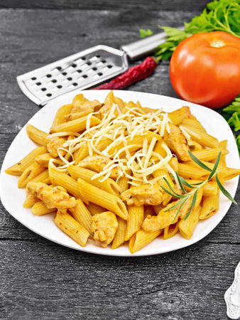Penne with chicken breast, tomato sauce and cheese in a plate, hot pepper, grater and fork on dark wooden board backgroundの写真素材