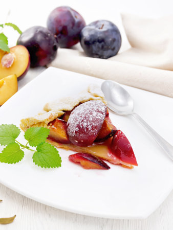Piece of sweet pie with plum, sugar, cardamom in a plate, sprigs of green mint, a kitchen towel on white wooden board backgroundの写真素材