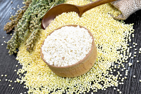 Millet flakes in a bowl, groats in a spoon, on a table and in burlap sack, stalks with ears of millet on dark wooden board backgroundの写真素材