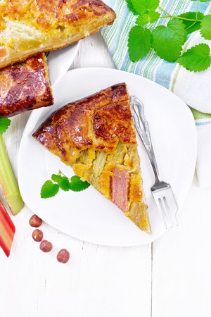 A piece of tart with rhubarb and nut cream in a plate, rhubarb stalks and nuts, a towel and mint on wooden board background from aboveの写真素材