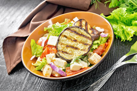 Salad of fried eggplant, fresh tomato, lettuce with red onion and salted feta cheese, seasoned with vegetable oil and soy sauce in a plate, towel, fork and parsley on wooden board backgroundの写真素材