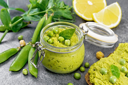 Hummus from green peas and chickpeas in a glass jar, pea pods, lemon and a sandwich on granite countertop backgroundの写真素材